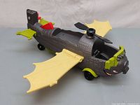 Side angle of the Madagascar 3 Crash Landing Plane toy showing yellow bat-like wings, detailed grey body, green and black accents, and a propeller on top.