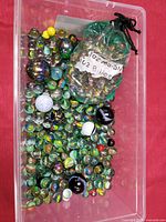 Top view of clear plastic bin filled with various vintage marbles, a green drawstring bag marked '102 Marbles' contains more marbles