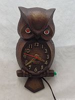 Front view of an electric owl-shaped clock with dark brown textured carved wood-like material, orange eyes, and an electric cord attached.