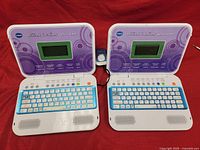 Pair of two purple and white Vtech Brilliant Creations Genius Junior beginner laptops shown open, side by side, displaying keyboard and screen area.