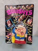 Front view of Squishys Commando squeezable toy sealed in original packaging showing caricatured character with red glasses and colorful outfit against black card with pink Squishys title and yellow burst graphic