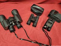 Overall view of three pairs of binoculars displayed on red fabric, showing various sizes and carrying cases.