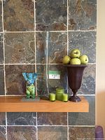 Wooden mantle with stone tile wall behind, showing the dark pedestal vase filled with 12 artificial green apples, next to a tall clear glass cylindrical vase, three small green candles, and a blue-green dog figurine on a gray base.