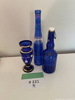Three blue glass pieces including a medium-sized hand-painted cobalt blue vase with gold accents, a tall slender gradient blue bottle, and a blue glass bottle with flip-top closure placed against a plain wall