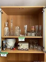 Glassware and barware items inside a wooden cabinet. Top shelf shows glass decanters, beer steins, and pilsner glasses. Bottom shelf has ice bucket, cocktail shaker, box of glasses, and stemmed glasses.