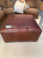 Front view of a large brown leather ottoman with square stitch pattern on top. Shows texture and light wear.