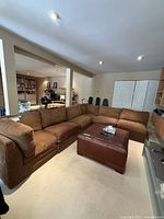 Six-piece brown modular sectional sofa arranged in living room area with beige carpet and light walls. Ottoman in front is not included in lot.