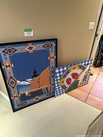 Photo showing two framed canvas art prints leaning against a beige wall on a light carpeted floor. One print with southwestern adobe design and geometric border; the other with still life on checkered background.
