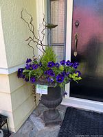 Full view of large metal urn planter with purple pansy flowers and attached decorative bunny water dish, placed outside next to house door.