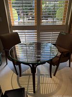 Front view of the round bistro table with glass top and rattan base, showing the table set against a window with chairs around it (chairs not included).