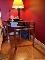 Side angle view of one brass and glass end table with lamp on top against red wall showing brass frame and glass surface