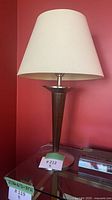 Retro style table lamp with a white tapered fabric shade on a dark brown tapered wooden base and metal round stand, placed on glass table.