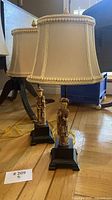 Pair of vintage Asian-style table lamps with carved figures on black pedestal bases and beige fabric shades with braided trim, photographed on a hardwood floor with surrounding furniture.