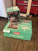Photo shows vintage Coleman green propane camp stove box with bilingual labeling and Coleman Insta-Lite propane lantern box on top.