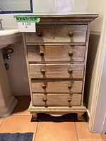 Front view of vintage solid wood 5-drawer dresser with glass top and brass handles, next to a pedestal sink on terracotta tile floor.