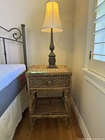 Wicker nightstand beside bed with lamp on top and window with white blinds in background