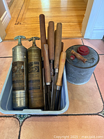 Photo showing two vintage brass pump fire extinguishers, multiple wood-handled chisels in a plastic bin, and a metal canister with red lid on floor.