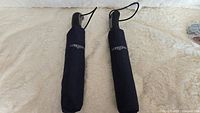 Two black compact umbrellas side by side on a soft beige surface with 'Longines' logos visible on protective covers.