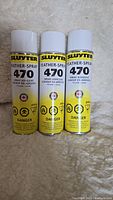 Front view of three new cans of Slyter Feather-Spray 470 yellow spray adhesive on a light carpet.