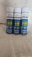 Three cans of Auto-Brite Premium Glass Cleaner standing upright on a light-colored fabric surface.