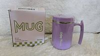 Photo showing the purple insulated mug next to its box labeled 'MUG' and '500ML'. Mug has a handle, lid, and 'Enjoy Time' print.