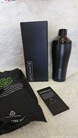 Matte black Brumate cocktail shaker with black box, instruction booklet, and black carrying bag on white surface