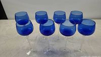 Photo of 8 blue wine glasses with clear twisted stems arranged on a table.
