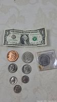 Overall lot showing one dollar bill, seven silver coins, one copper coin, and one silver dollar coin in plastic sleeve