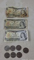 Three circulated Canadian one dollar bills showing wear, creases, and discoloration, laid above seven Canadian silver dollar coins.