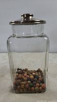 Front view of clear square-shaped early glass container filled with multiple small, round colored clay marbles, topped with a metal lid showing wear.