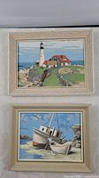 Photo of both framed paintings showing coastal scenes, lighthouse picture on top, boats picture on bottom.