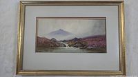 Framed signed watercolour painting titled "The Dart," showing landscape scene with river, rocks, purple plants, and distant mountains, in gold-toned frame with cream mat.