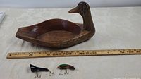 Full view of the wooden loon-shaped nut bowl with a ruler to show size, and both vintage fishing lures below it on a white tablecloth.