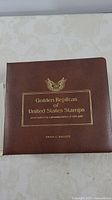 Brown collector's album with gold embossed text 'Golden Replicas of United States Stamps proof replicas on a glowing surface of 22kt gold' and Grace G. Jaggett's name.