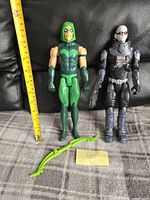 Mr. Freeze and Green Arrow action figures displayed side by side with measuring tape showing approximately 12 inches tall. Both figures include their respective accessories placed in front.