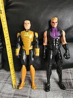 Two 12-inch action figures, Lex Luthor in black and yellow armor and Hawkeye in black and purple tactical gear, positioned side by side with a ruler showing height. Figures on a black background.