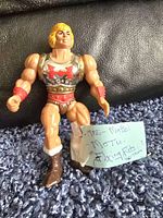 Front view of 1985 Mattel Flying Fists He-Man action figure standing on blue carpet with note card.
