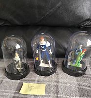 Three collectible figures under clear dome displays on black bases. From left: black dressed soldier/prisoner, blue and white superhero, pair of two figures in blue and green suits.