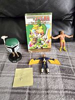 All four items pictured on a gray surface including the book, bendable Hulk Hogan figure, Mandalorian Funko Pop, and Max Steel figure.