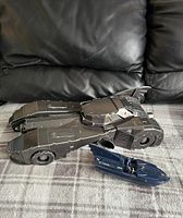 Retro 3D Batmobile model next to plastic Batboat on fabric surface with black couch background. Show size comparison and condition.