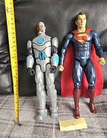 Front view of Cyborg and Superman action figures standing side by side with a yellow measuring tape showing height measurement around 12 inches