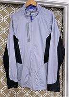 Front view of gray Adidas jacket with black accents hanging on hanger showing overall garment design and zippered chest pocket.