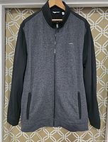 Front view of the men's Calvin Klein light jacket hanging on a wooden hanger on patterned background, showing full zip closure and side pockets.