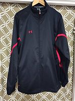 Front full view of the black Under Armour jacket with red accents hung on a hanger against a patterned background.