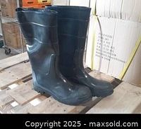 Pair of new men's black rubber boots standing upright on a wood pallet in front of cardboard boxes. The boots are around mid-calf height showing smooth black finish and rugged soles.