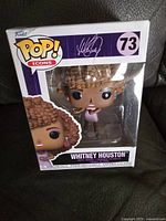 Photo of Whitney Houston Funko Pop figure in original packaging, front view showing figure and box design with purple accents, signature, and number 73.