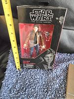 Front view of Han Solo action figure in original box with measuring tape showing height around 6 inches. Box has sticker residue on the top right corner.