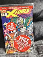 Front view of sealed X-Force #1 comic book with red and orange cover art and promotional stickers for the included trading card.