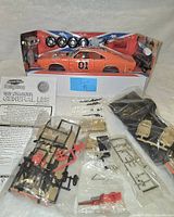 Die-cast 1969 Charger General Lee model in opened display packaging surrounded by sealed bags of parts and instruction sheet, parts include black frame components, beige seats, red and black plastic pieces.