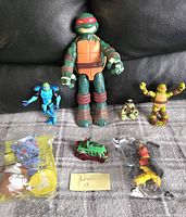 Photo of Teenage Mutant Ninja Turtles action figures including a large Raphael figure, smaller turtle figures, a blue robotic figure, a red vehicle, and two accessory packs in plastic.